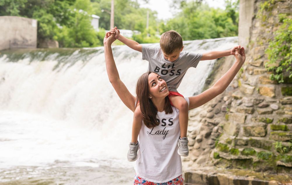 A child rides on someone's shoulders near a scenic waterfall and stone wall on a bright summer day.