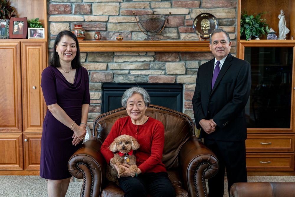 Three generations pose with a small dog in front of a stone fireplace in a cozy living room.