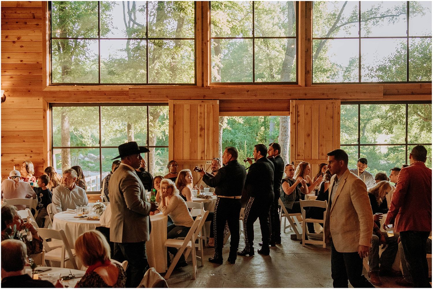 Wedding guests gathered in a sunlit wooden reception hall with large windows overlooking nature.