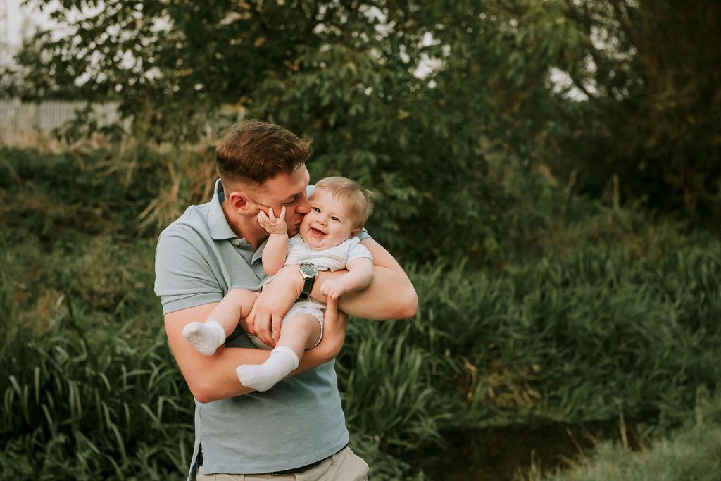 Father holding and playing with baby outdoors in natural setting during sunset.