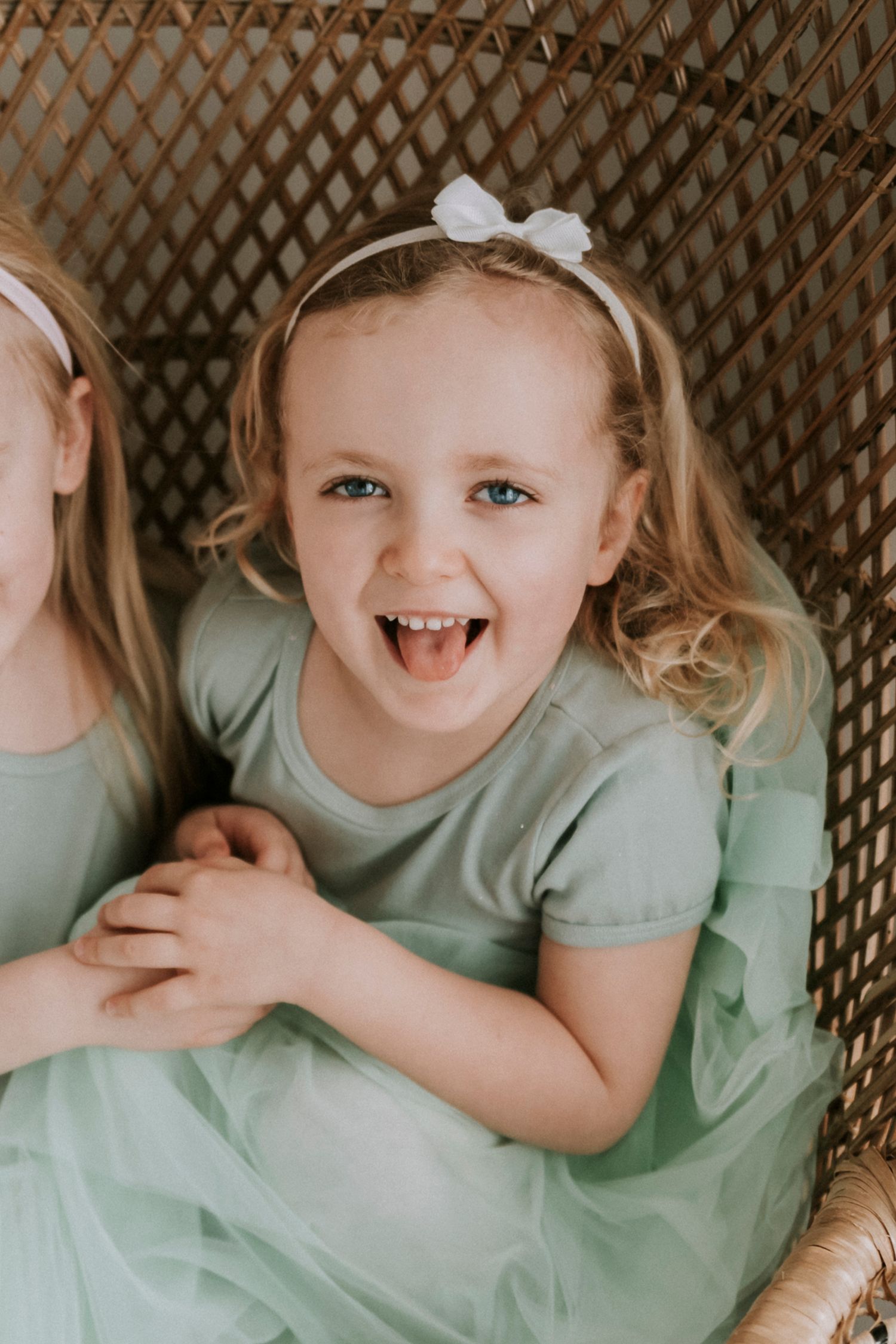Small child in mint green dress sitting in wicker chair with joyful expression.