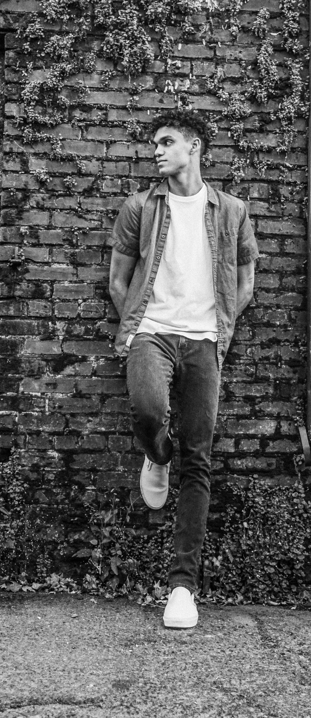Moody black and white portrait of a person leaning against a brick wall covered in vines.