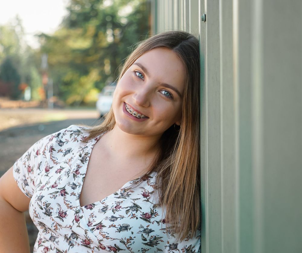 Portrait of someone in floral print top smiling warmly while leaning against wall outdoors.