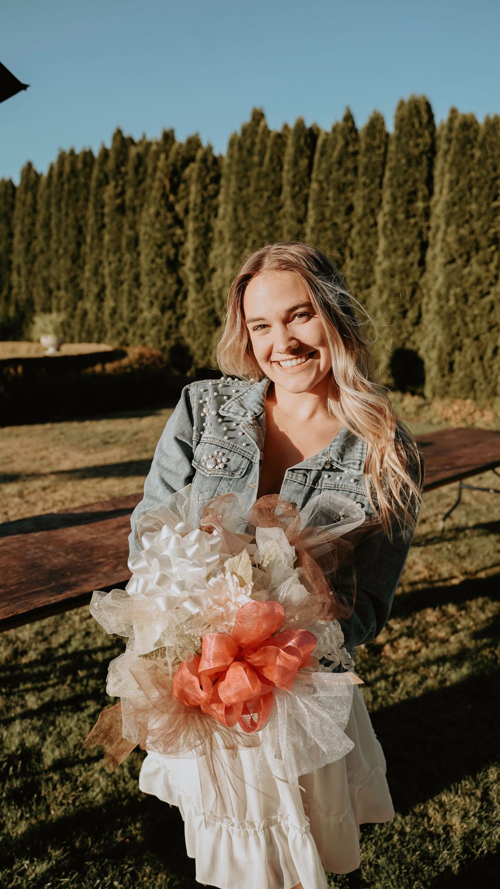 Portrait against cypress trees showing a white lace dress and denim jacket with decorative coral and white flowers.