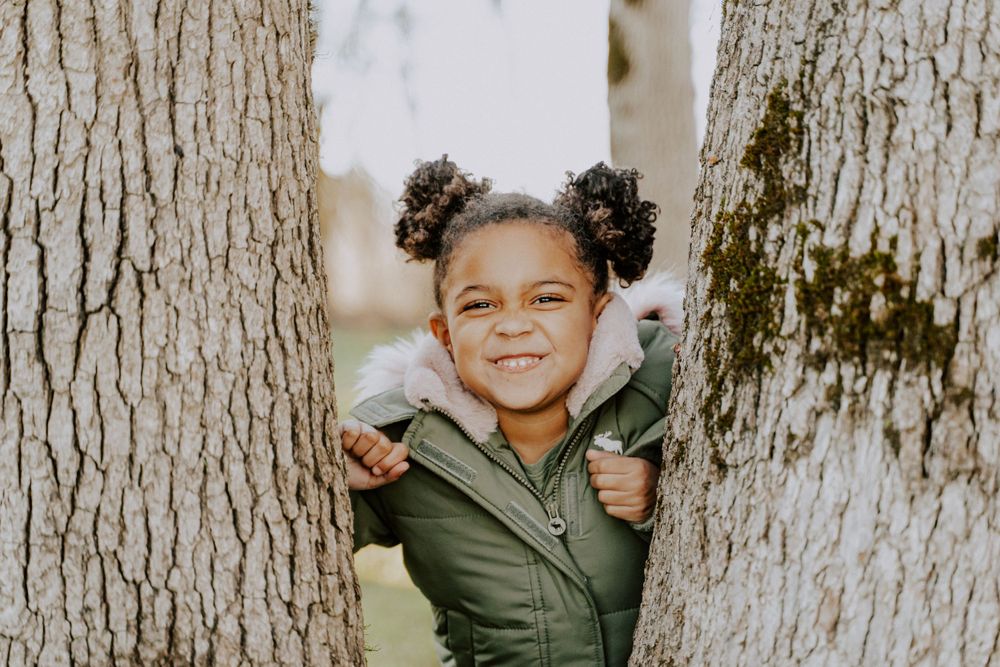 Joyful child peeks through tree trunks with hair styled in two buns.