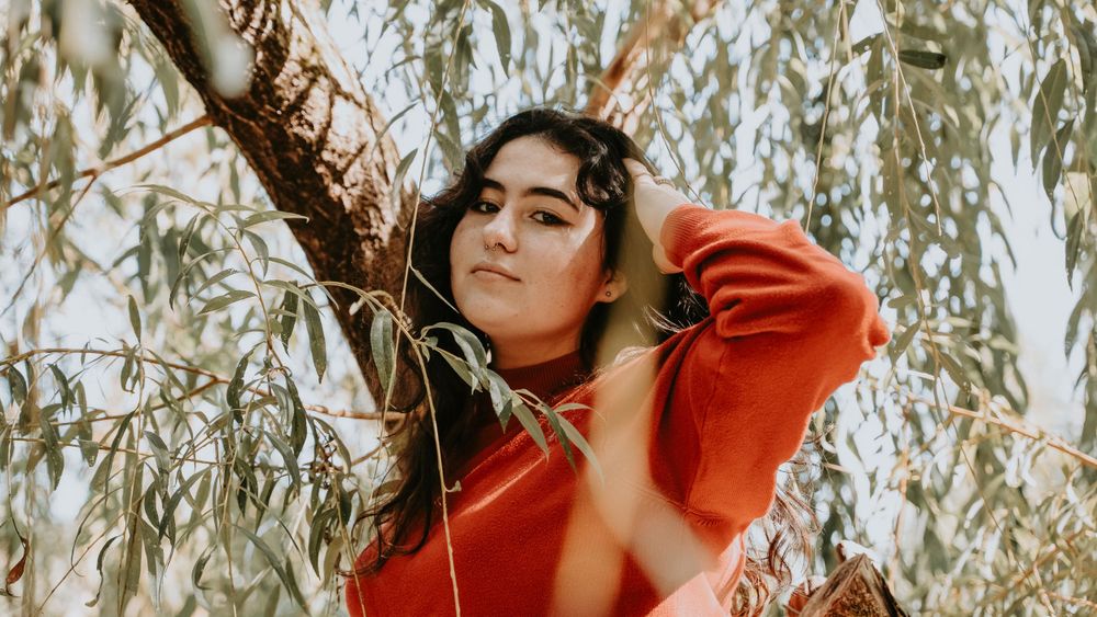 Person in red sweater posing dramatically under willow tree branches.