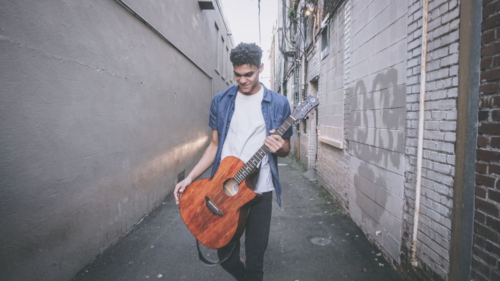Person holding an acoustic guitar in an urban alley wearing casual attire.