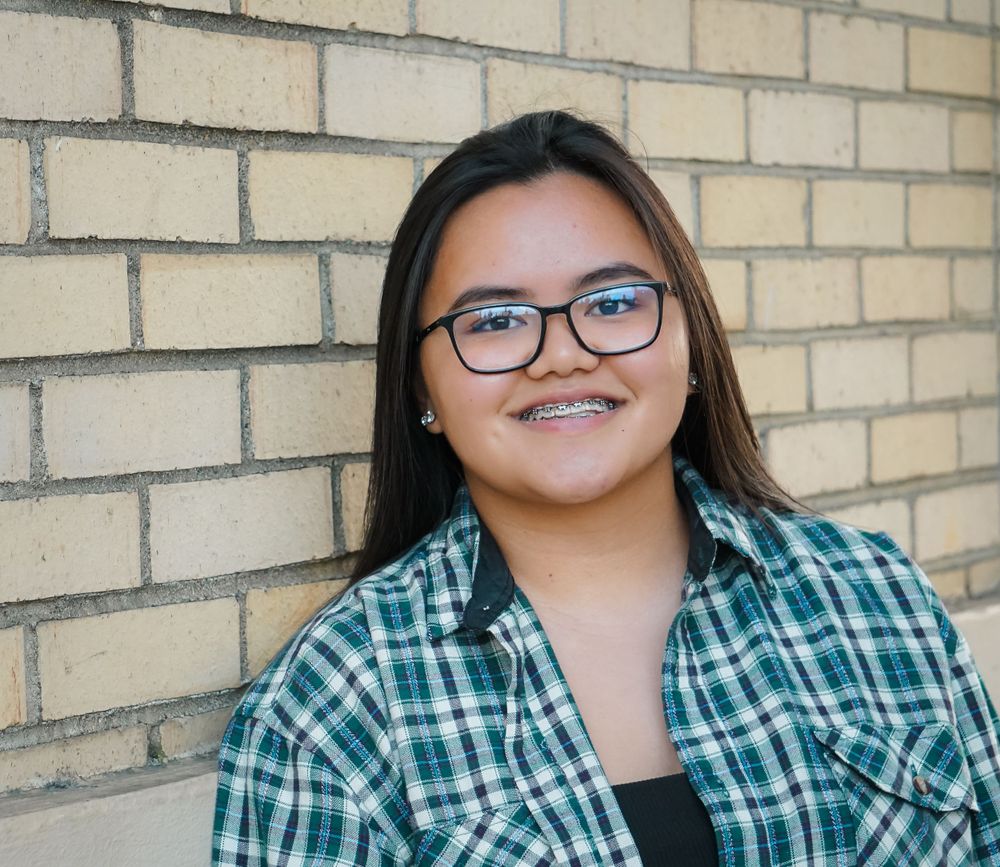 Portrait of someone in glasses and plaid shirt smiling against brick wall background.
