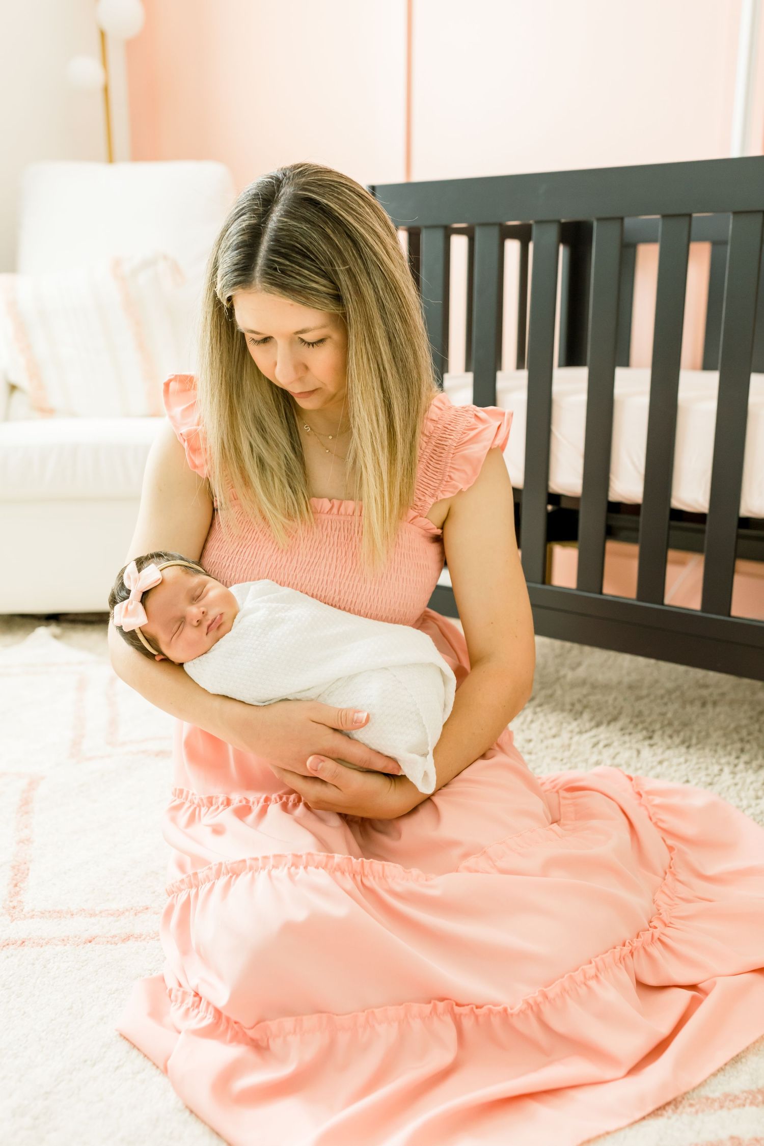 Nursery scene with caregiver in pink dress holding swaddled infant near dark crib.