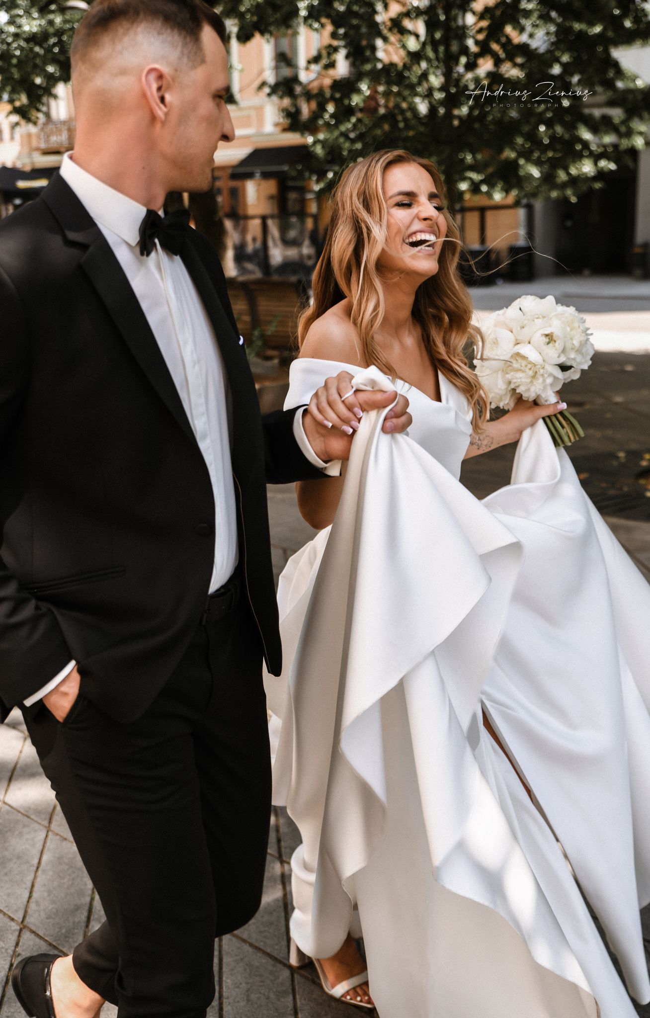 Couple sharing joyful moment outdoors, bride in elegant off-shoulder gown holding white bouquet.
