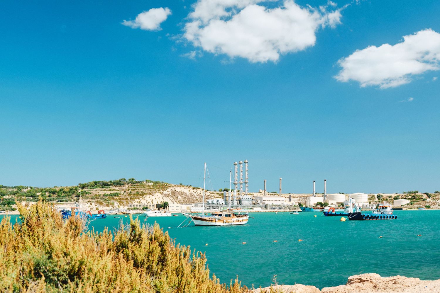 Scenic harbor view with boats and industrial structures against bright blue sky.
