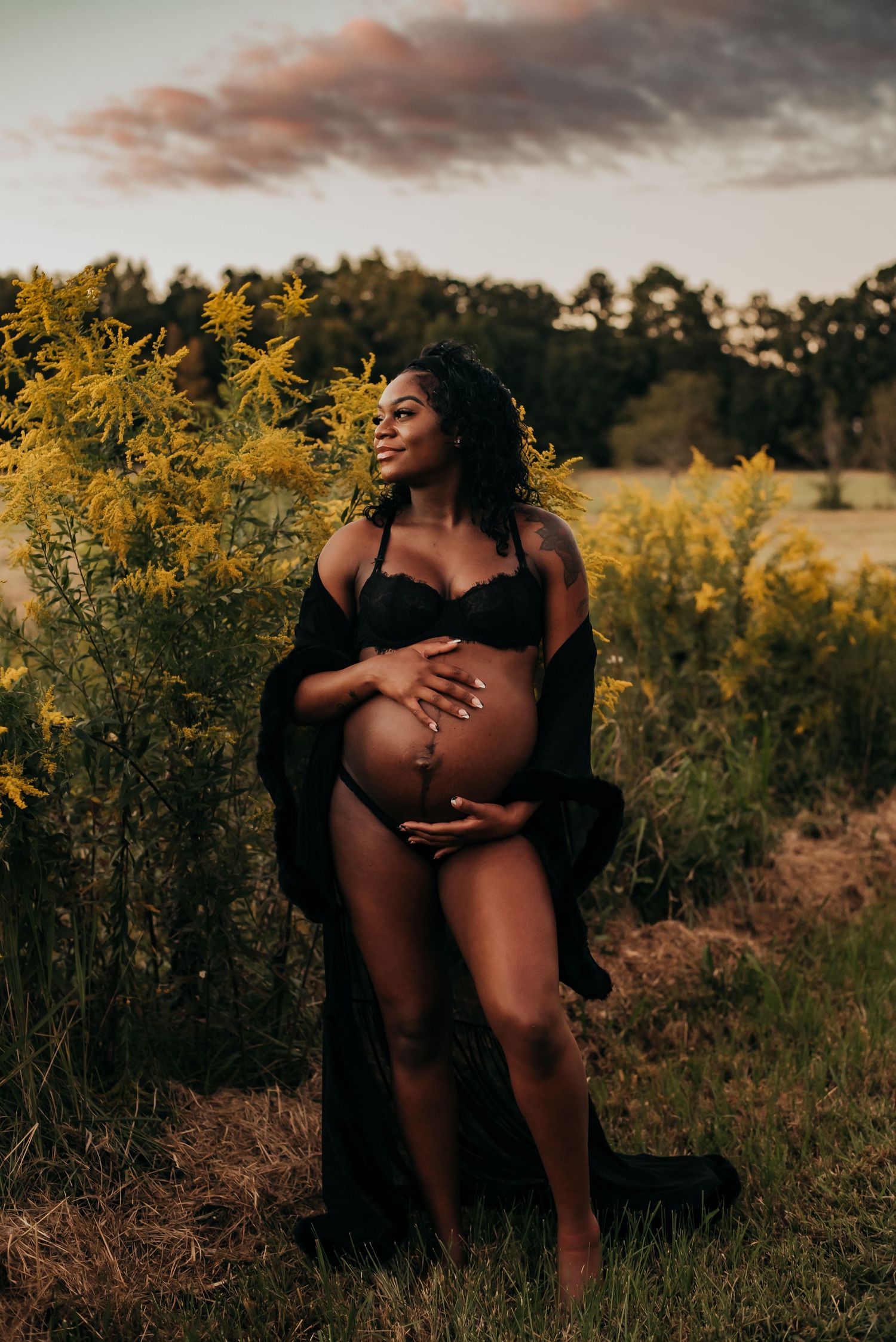 Pregnant woman in black lingerie posing near yellow wildflowers at sunset.