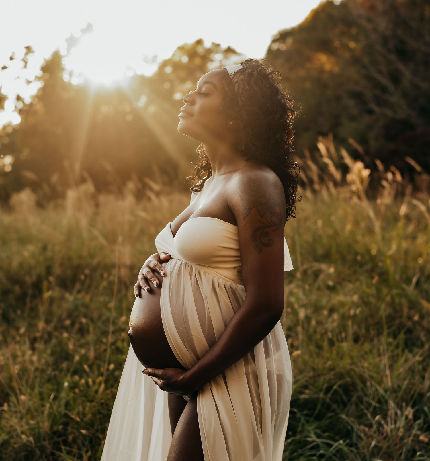 Pregnant woman in sheer cream gown holding belly in golden sunlit field.