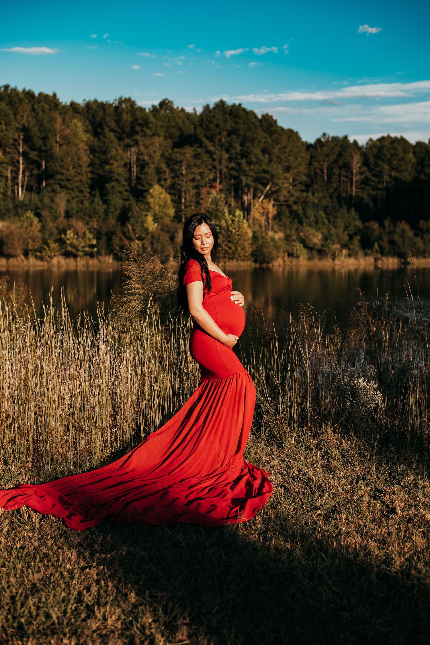 Pregnant woman in red mermaid gown posing near lake with forest backdrop.
