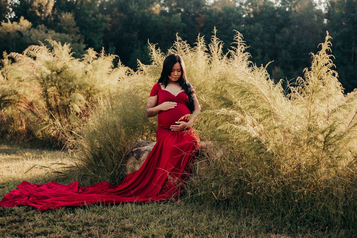 Pregnant woman in red gown sitting among tall ornamental grasses outdoors.