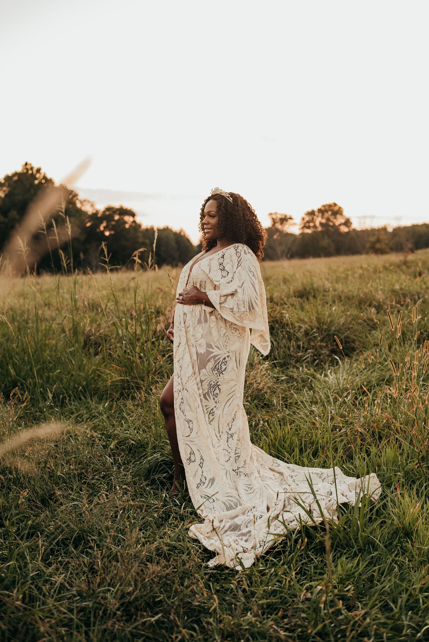 Pregnant woman in white lace maternity gown smiling in sunlit meadow.