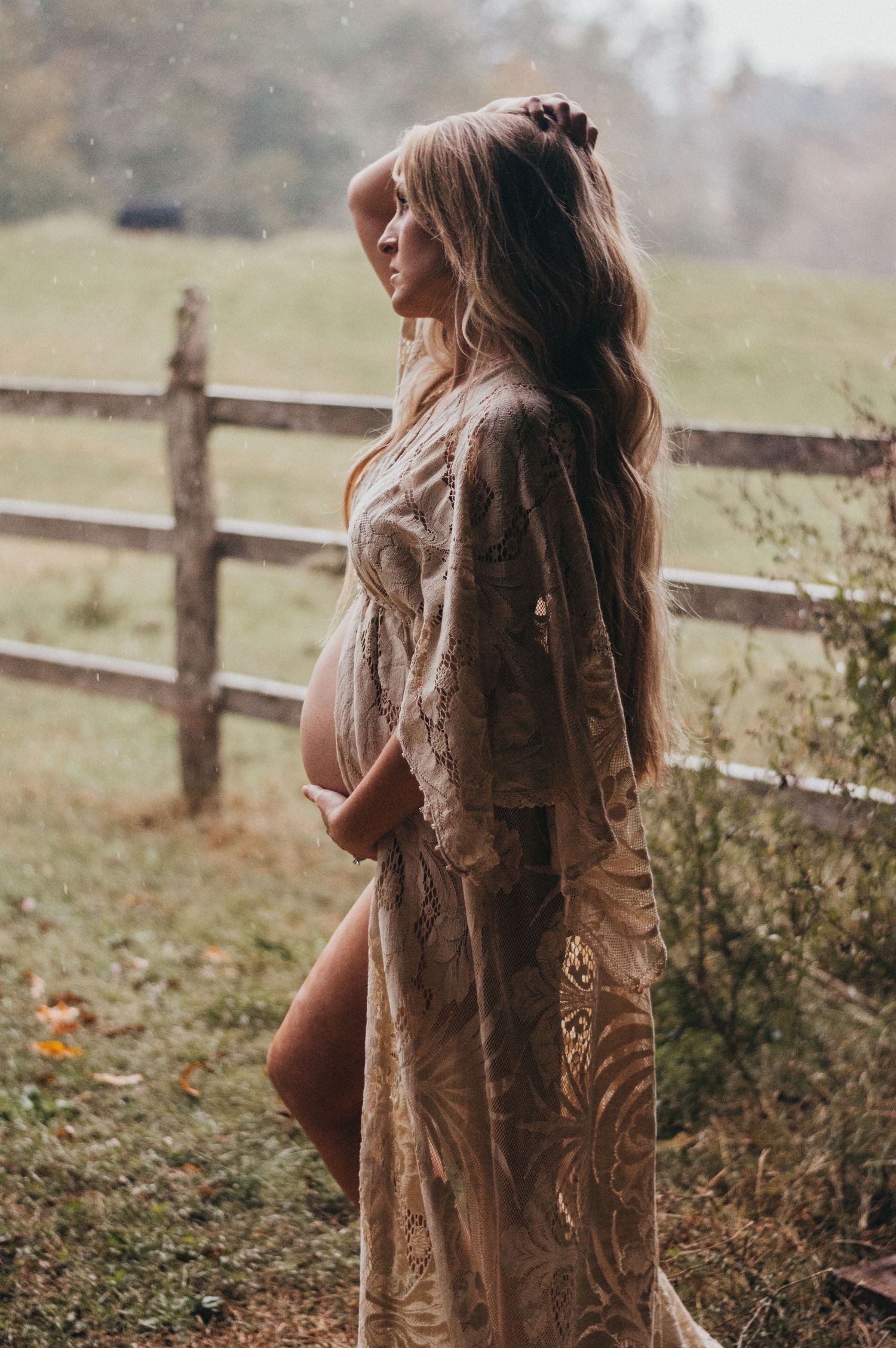 Pregnant woman in lace gown holds baby bump outdoors near rustic fence in misty field.
