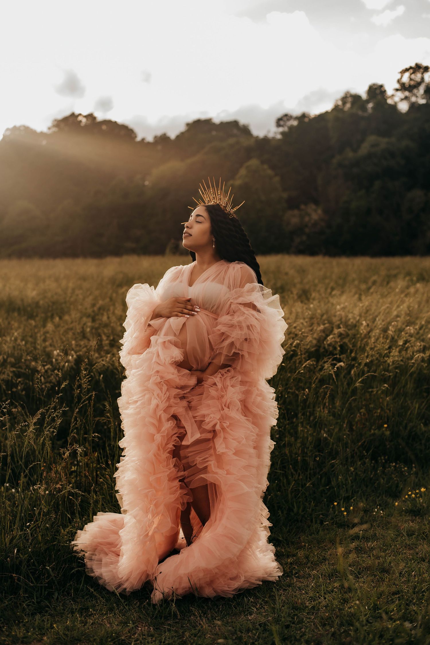 A pregnant woman in a voluminous pink tulle gown wears a gold crown in a sunlit meadow.