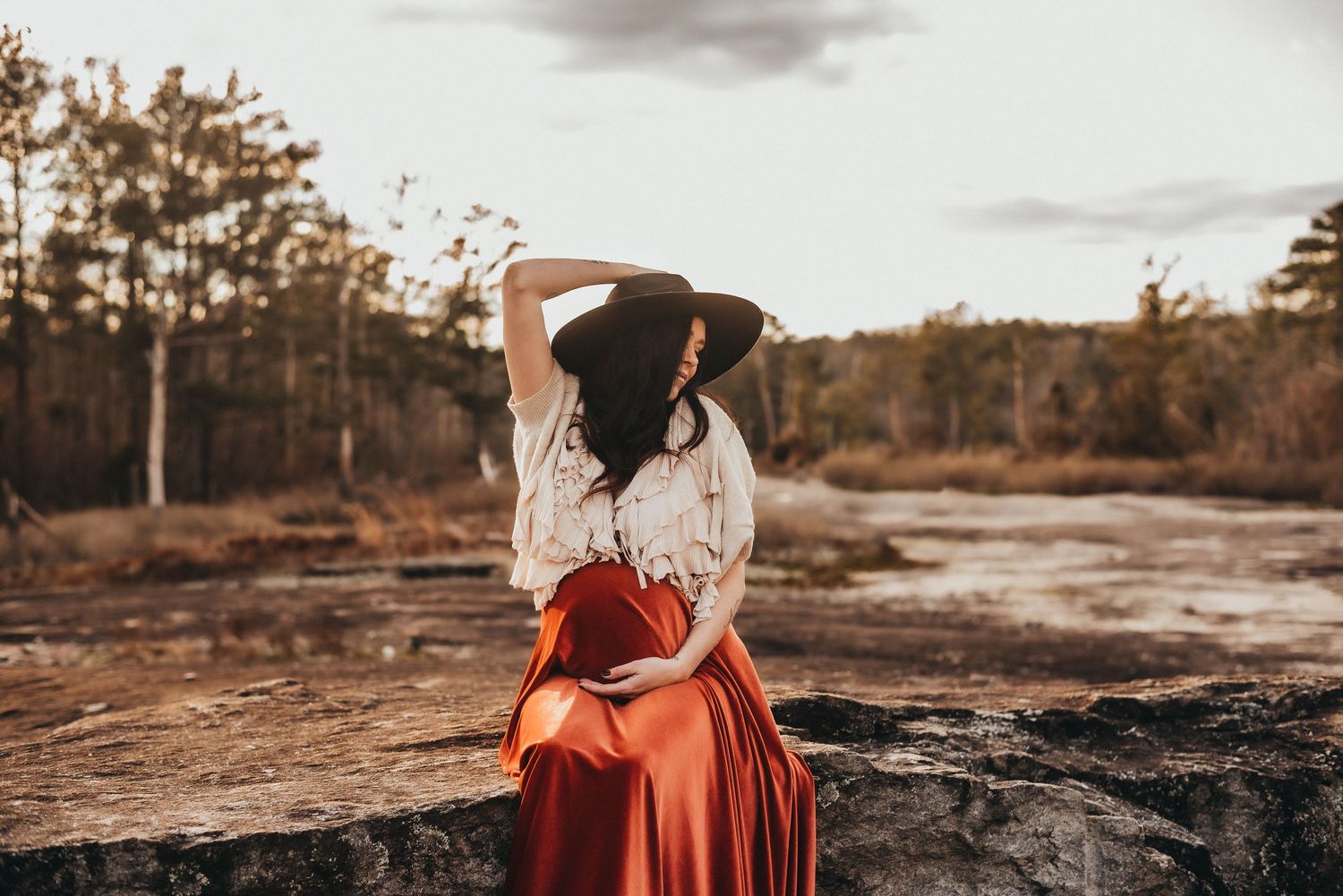 Pregnant woman in red skirt and wide-brim hat sitting on rocks at sunset.