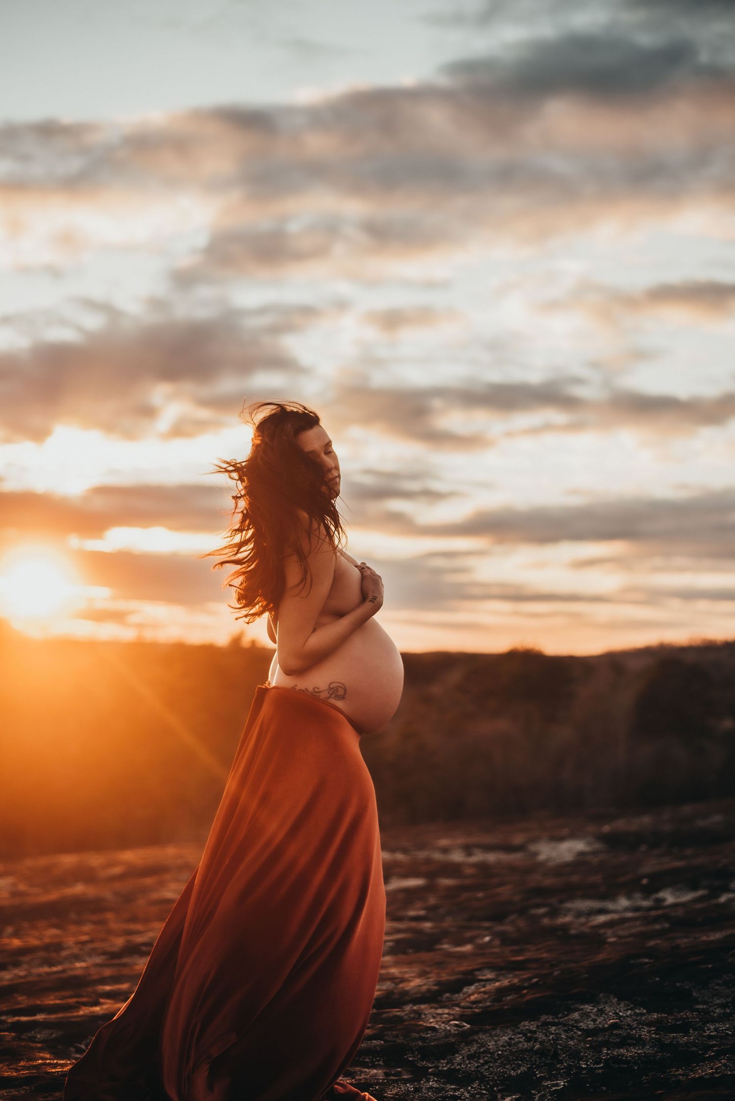Pregnant woman in red skirt cradling bump at dramatic golden sunset outdoors.