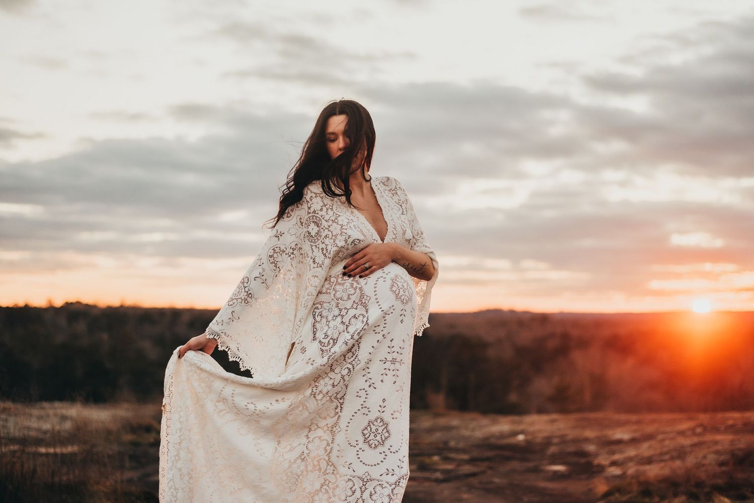 Pregnant woman in white lace boho dress posing at sunset in open field.