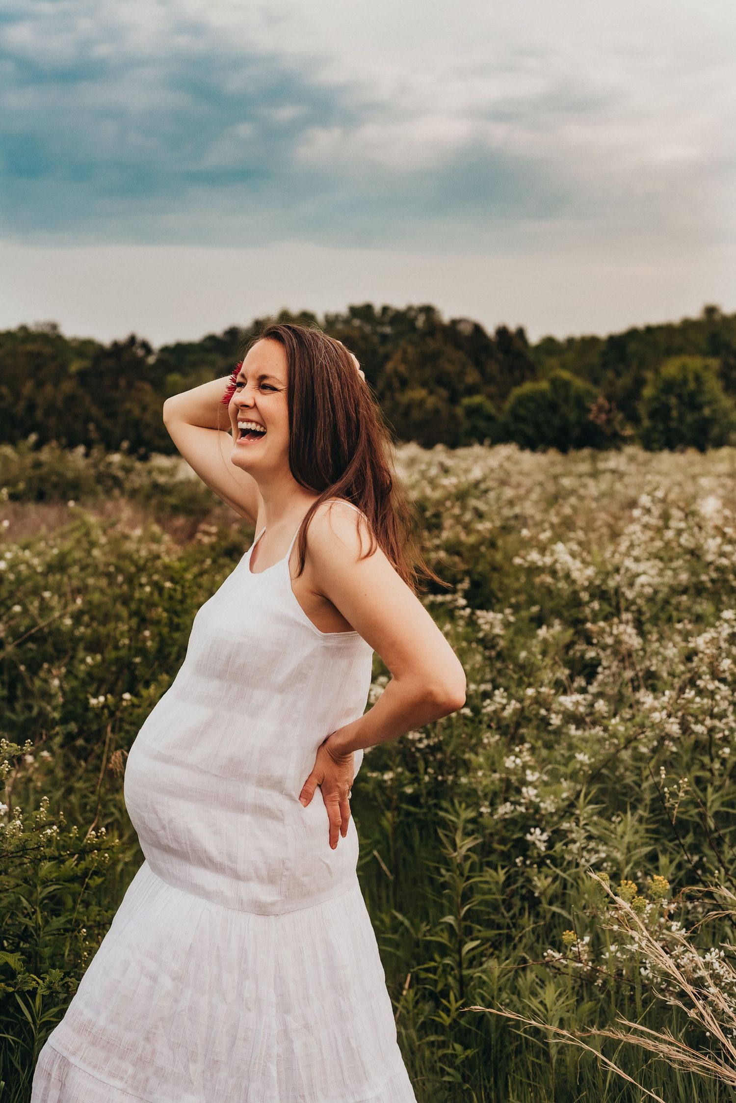 Laughing pregnant woman in white dress standing in a wildflower field.