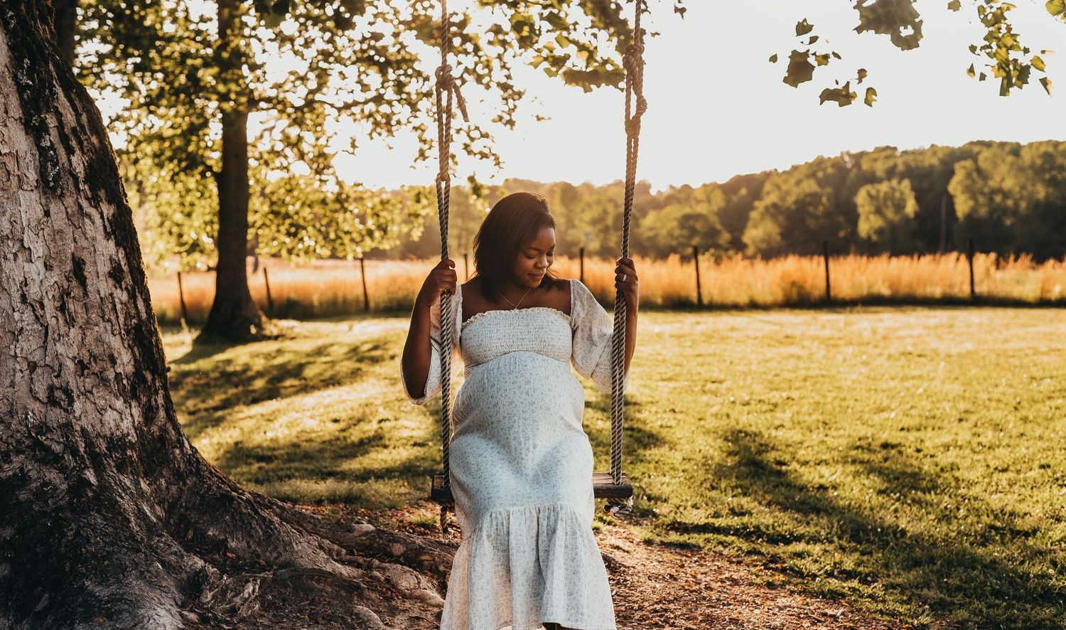 Pregnant woman in white dress sitting on tree swing in golden hour sunlight in lush green park.