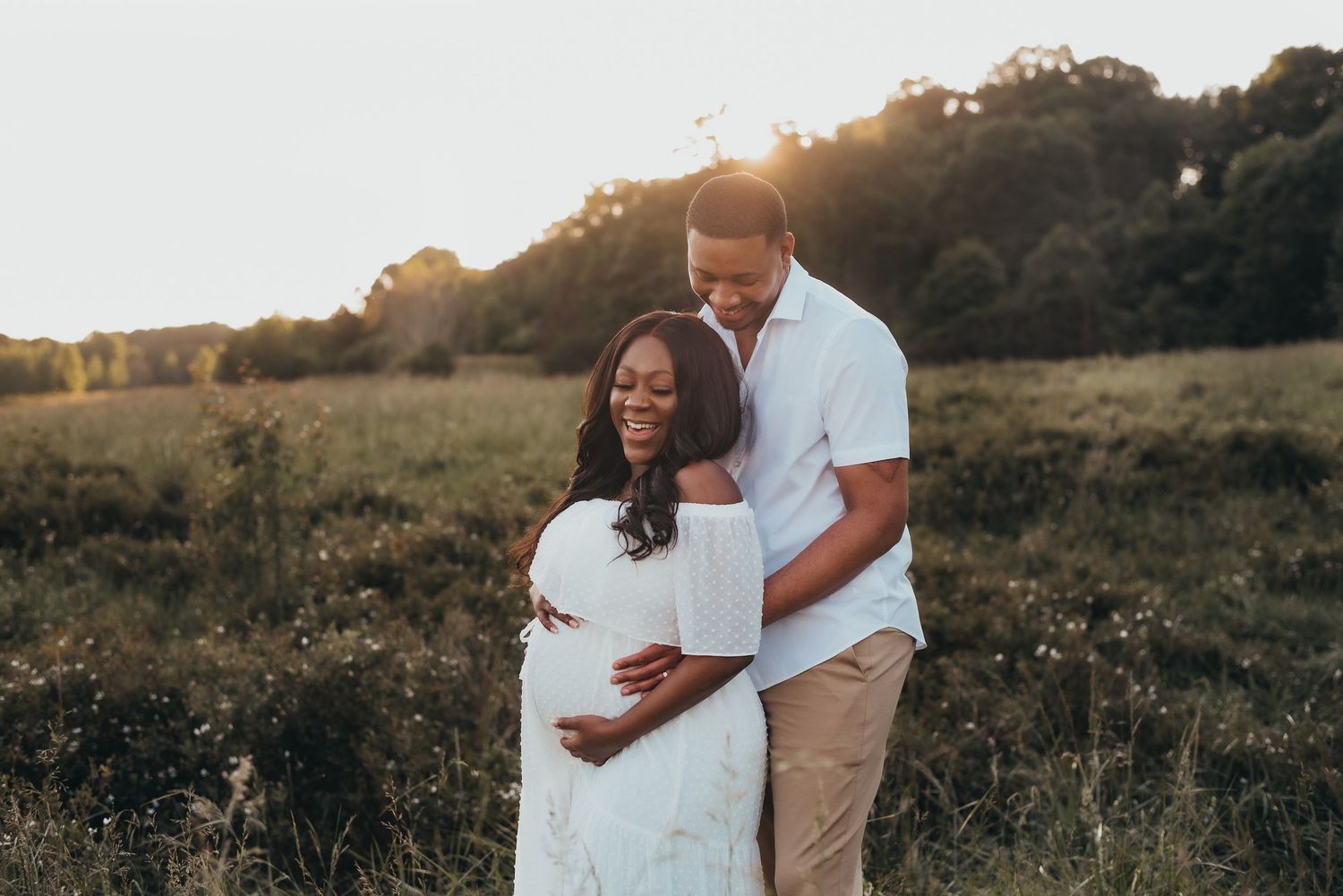 Pregnant Black couple embracing in a golden sunset field, woman in white dress holding baby bump.