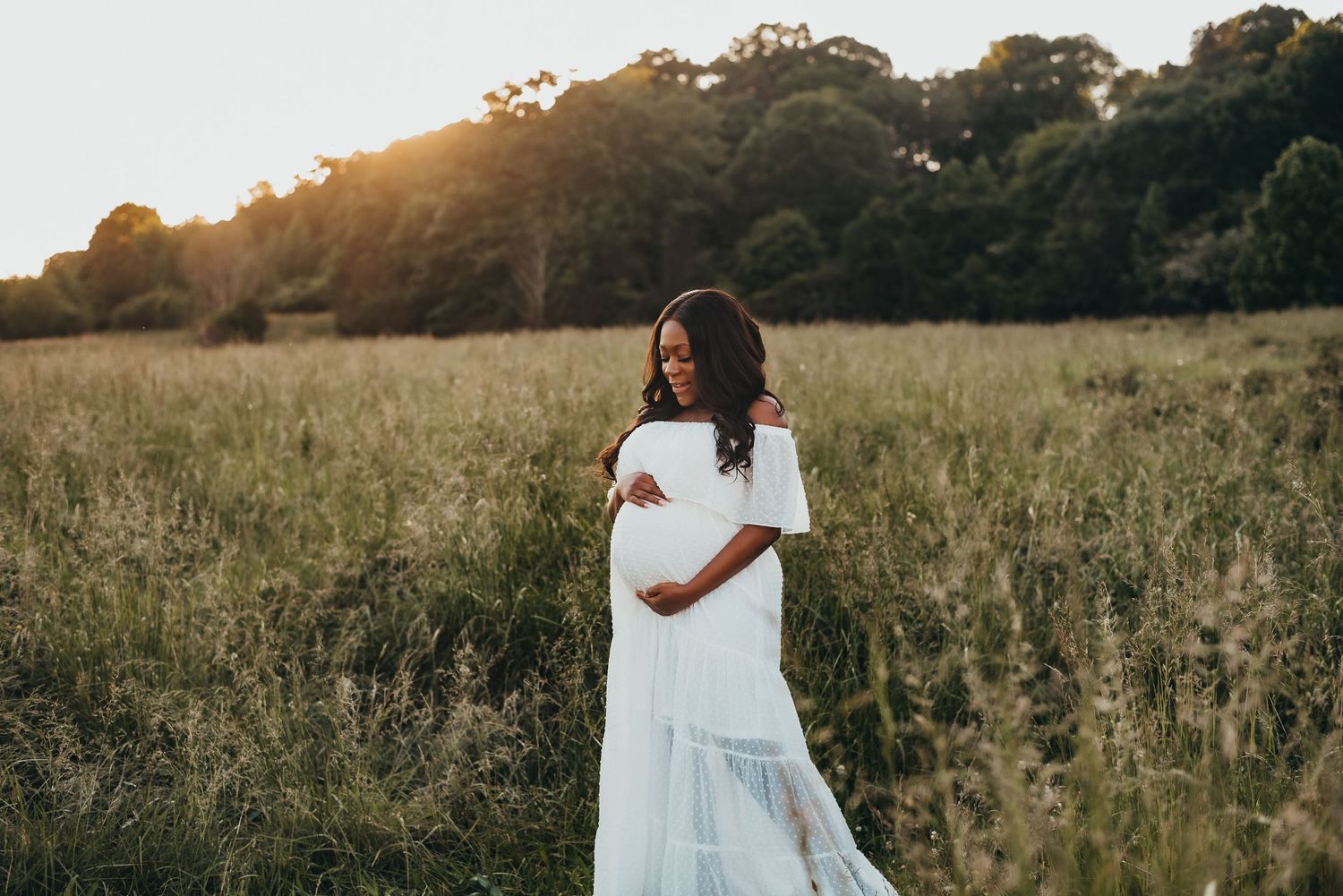 Beautiful pregnant Black woman in white off-shoulder gown standing in golden meadow at sunset.