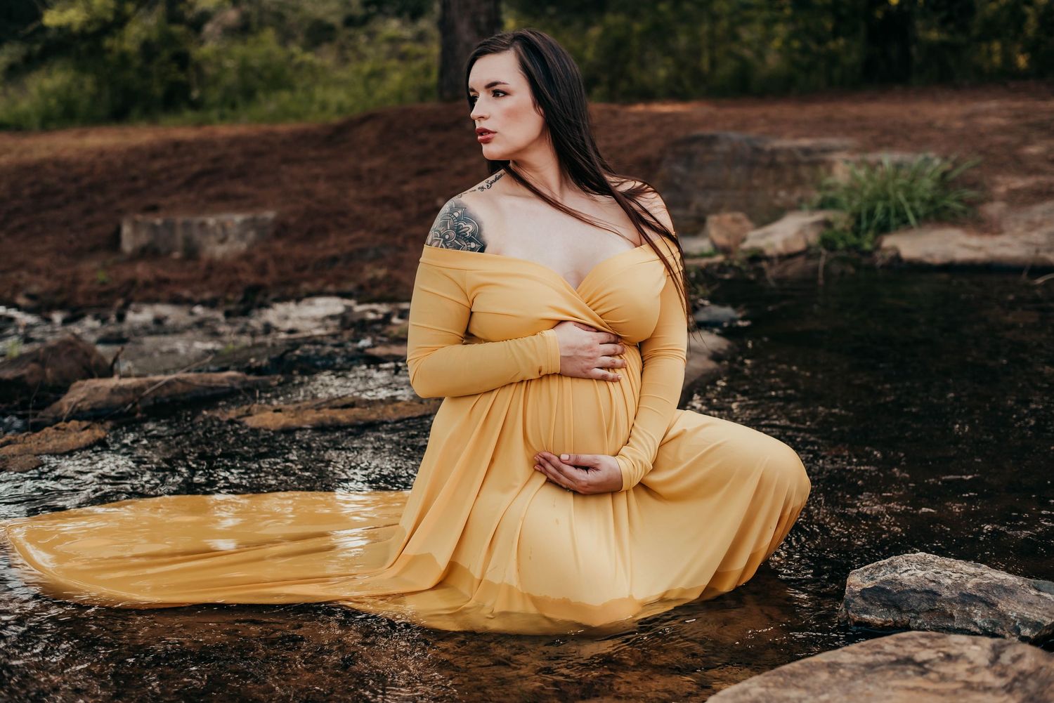 Pregnant woman in flowing yellow gown sitting gracefully on rocks beside a serene stream.