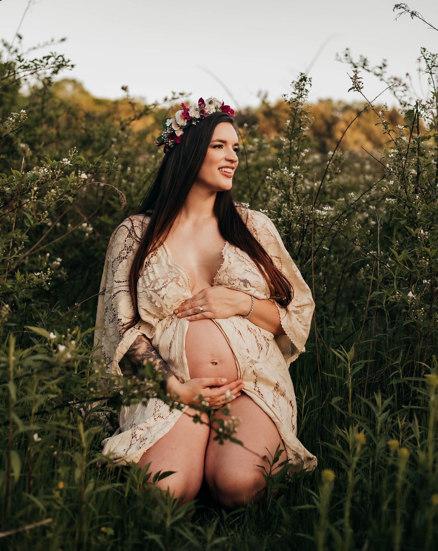 A smiling pregnant woman in lace robe and floral crown kneels among wildflowers at golden hour.