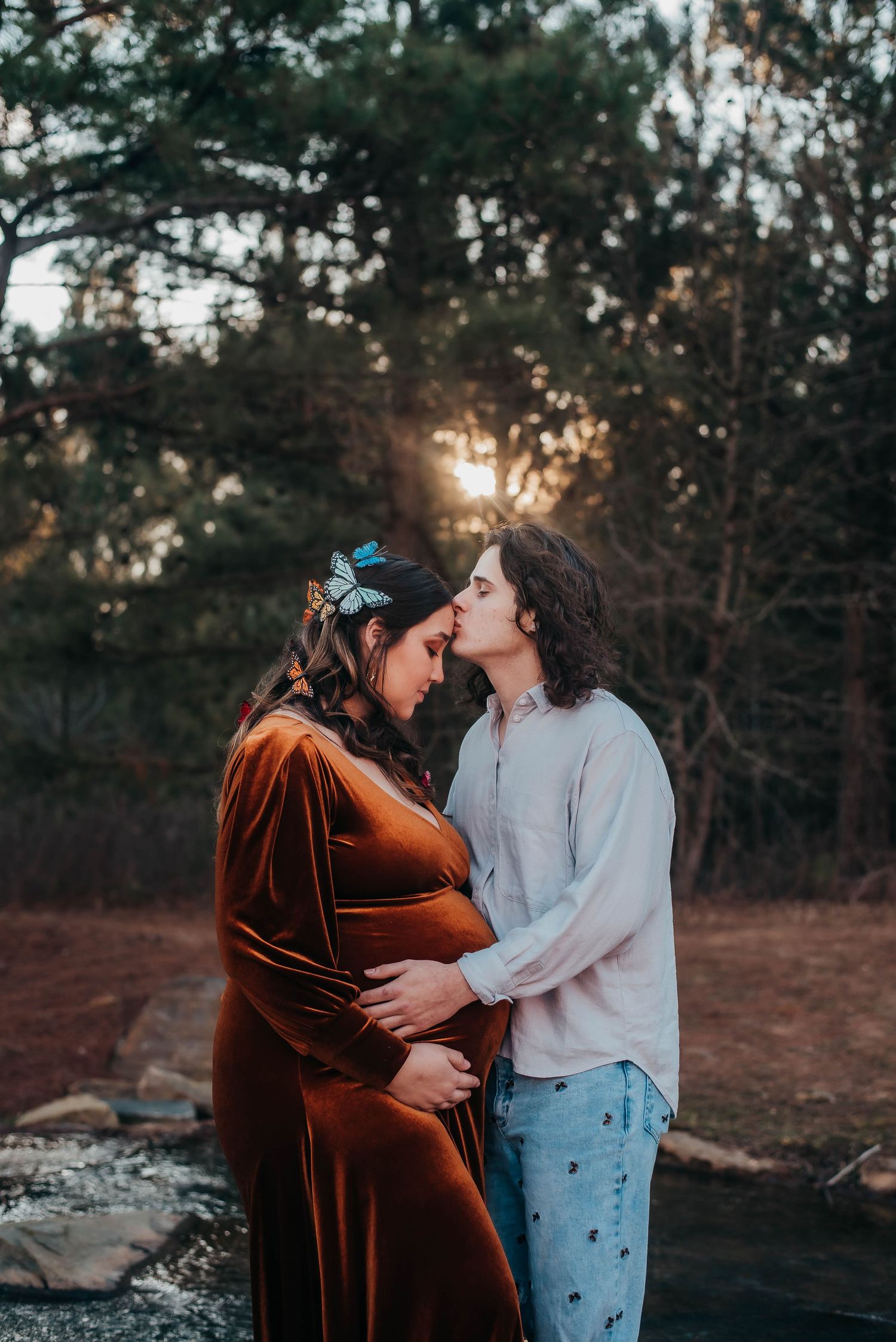 A couple shares an intimate kiss while cradling a baby bump at golden hour outdoors.