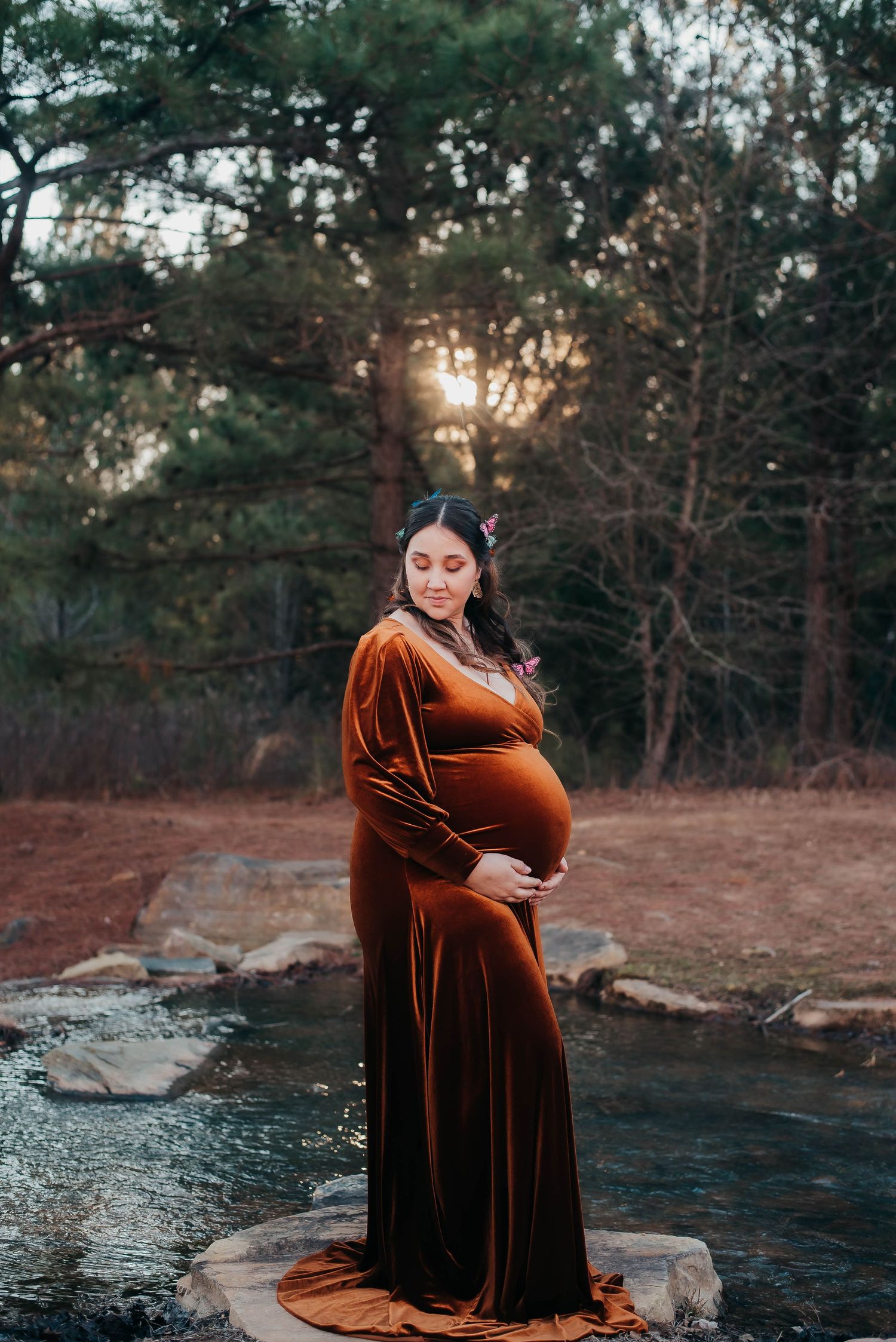 A pregnant woman in a rust velvet gown cradles her belly by a sunlit forest stream.