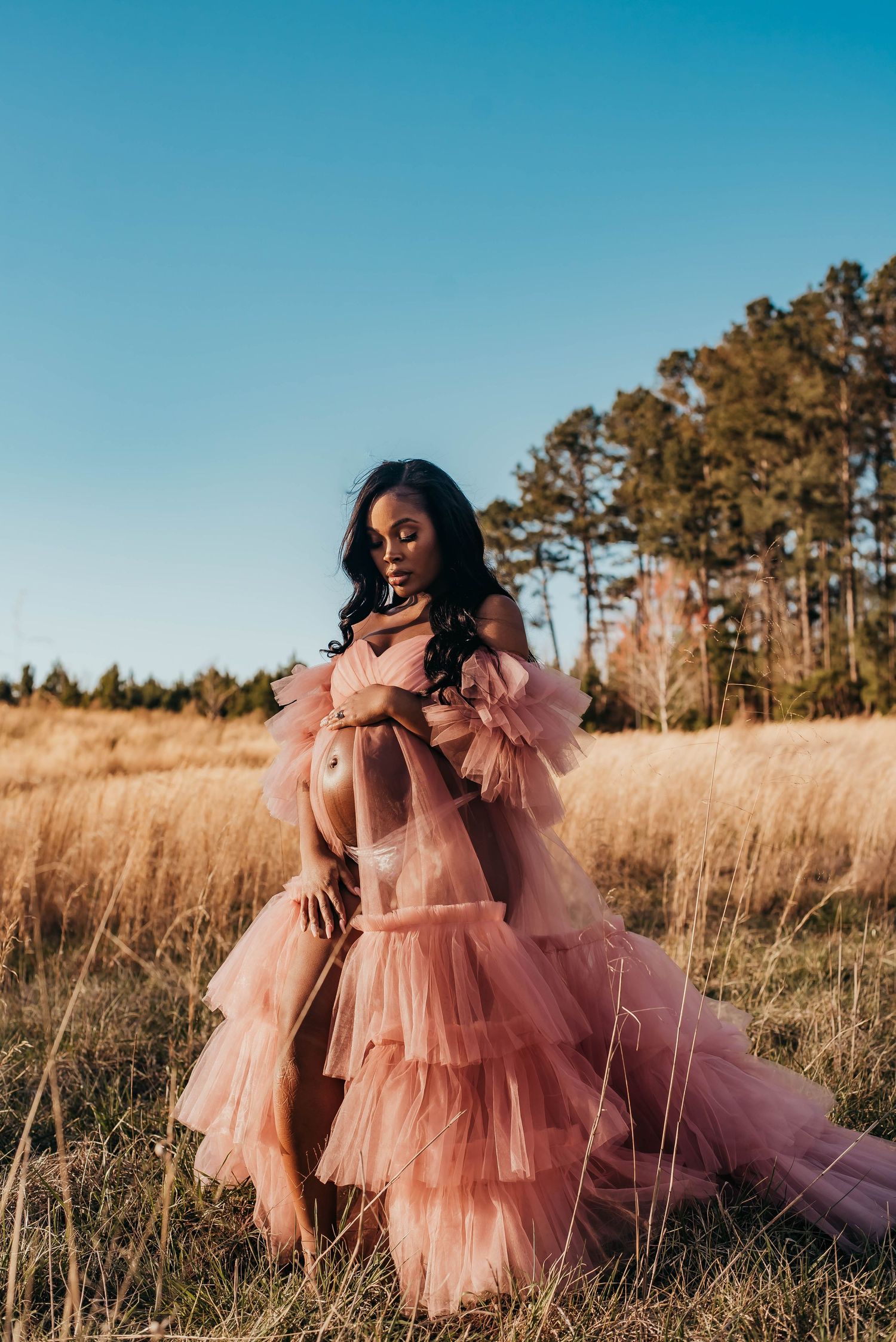 A pregnant woman poses in a flowing pink tulle gown in a golden field under a clear blue sky.