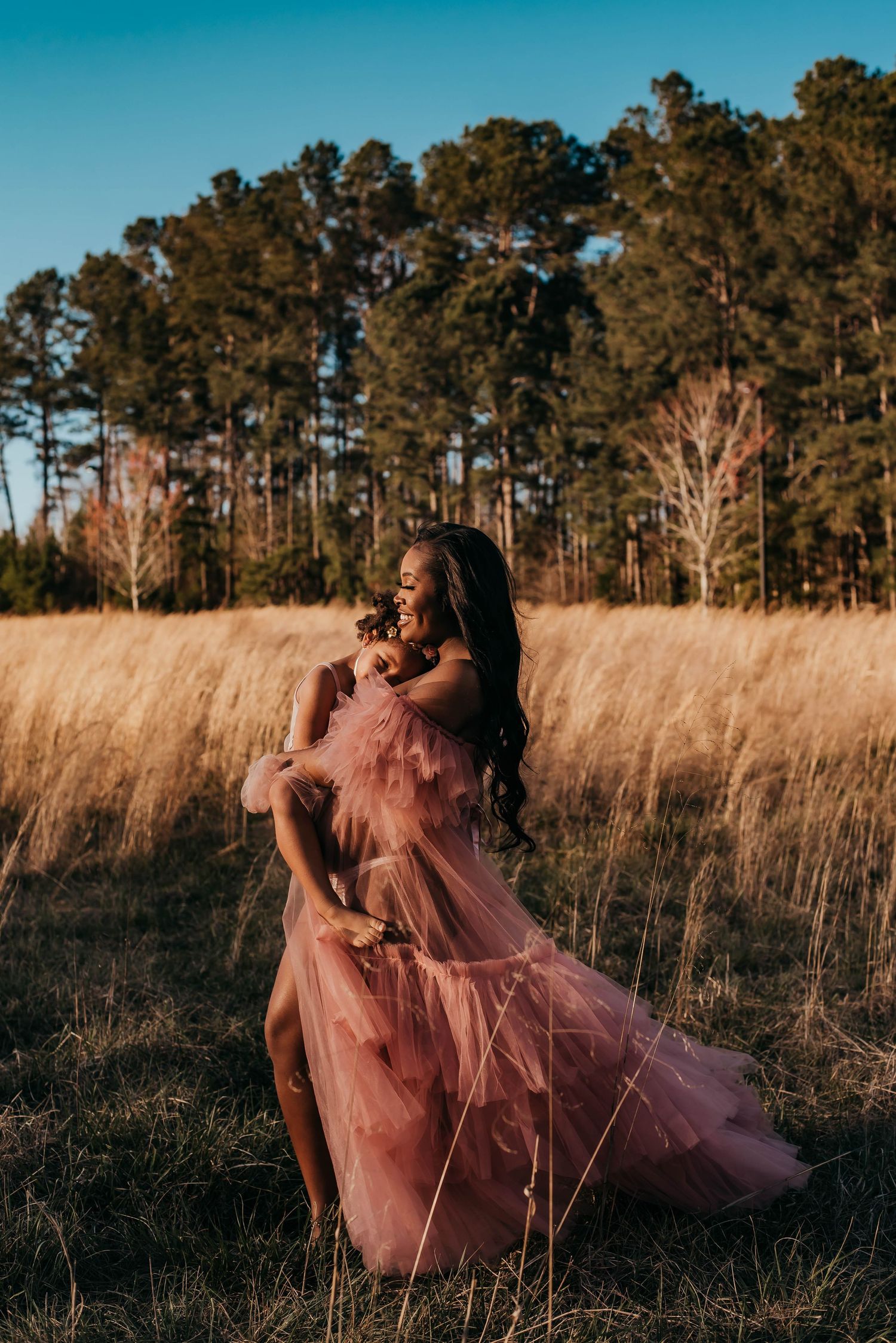 A pregnant woman in a layered pink tulle gown twirls gracefully in a sunlit golden grass field.