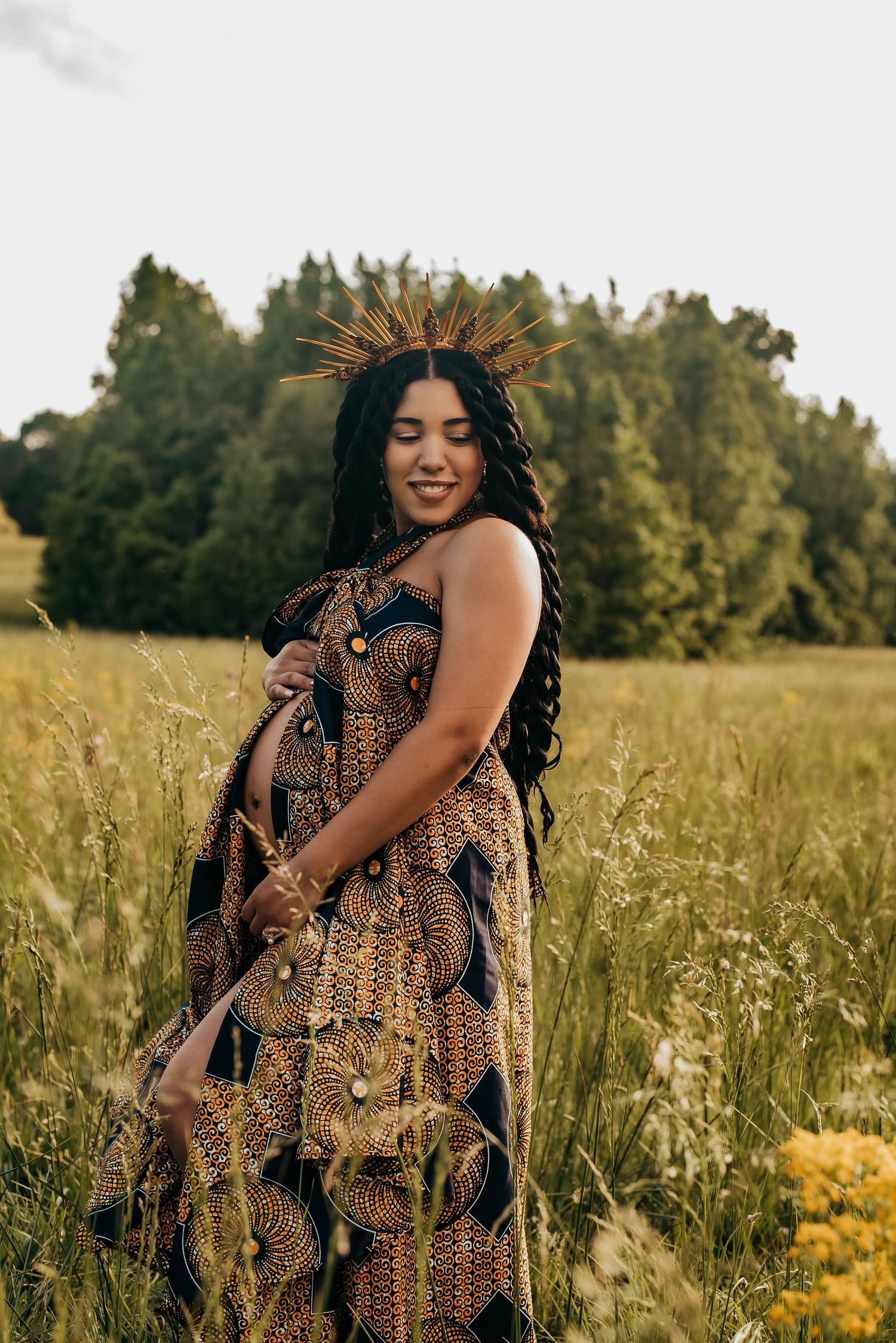 A pregnant woman in a gold African print dress and sunburst crown smiles in a wildflower field.