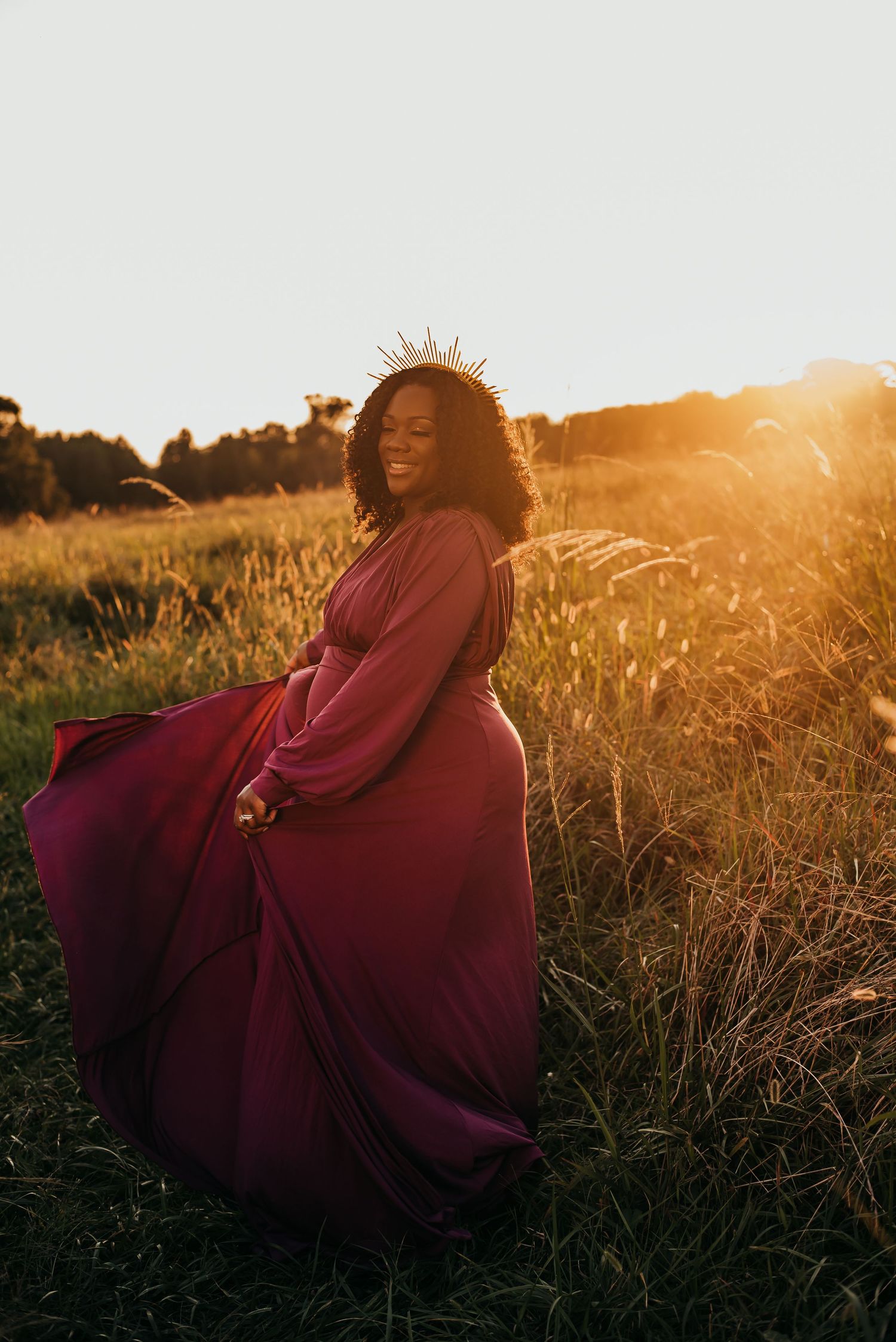 Pregnant woman in flowing burgundy gown wearing crown twirling in golden field.