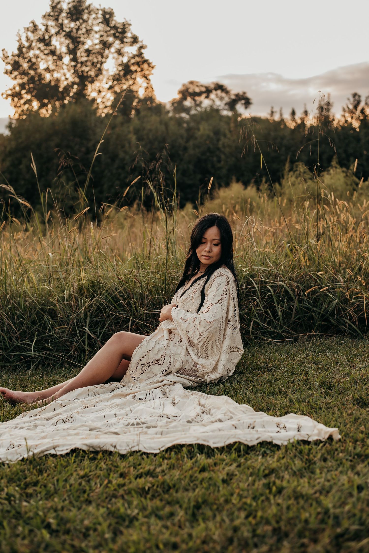 Pregnant woman in cream lace robe sitting on blanket in grassy field.