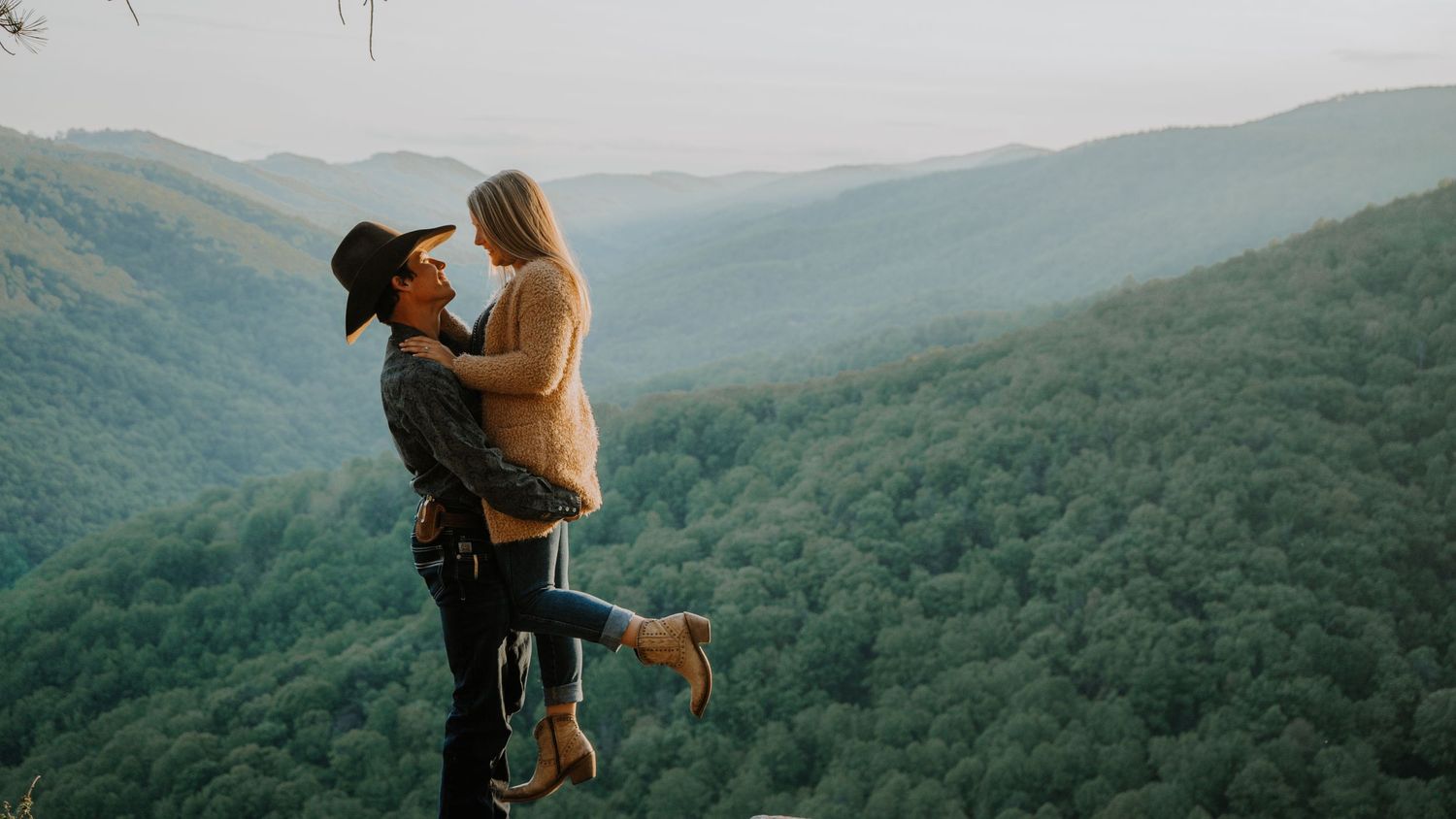 Two people embrace on mountain overlook during sunset with rolling hills in background.