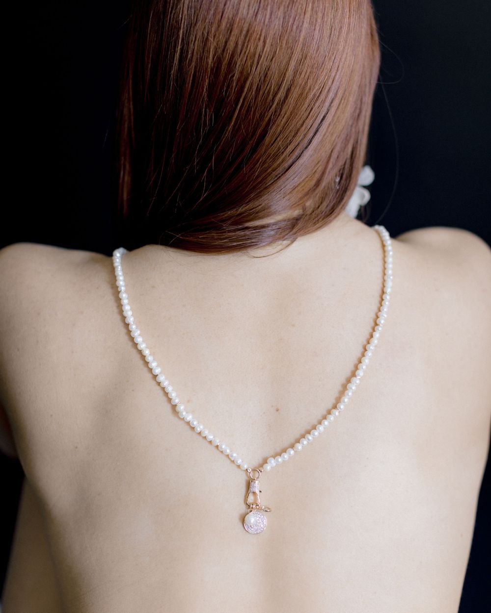 A delicate pearl necklace adorns a bare back against a dark background, with long brown hair falling to one side.