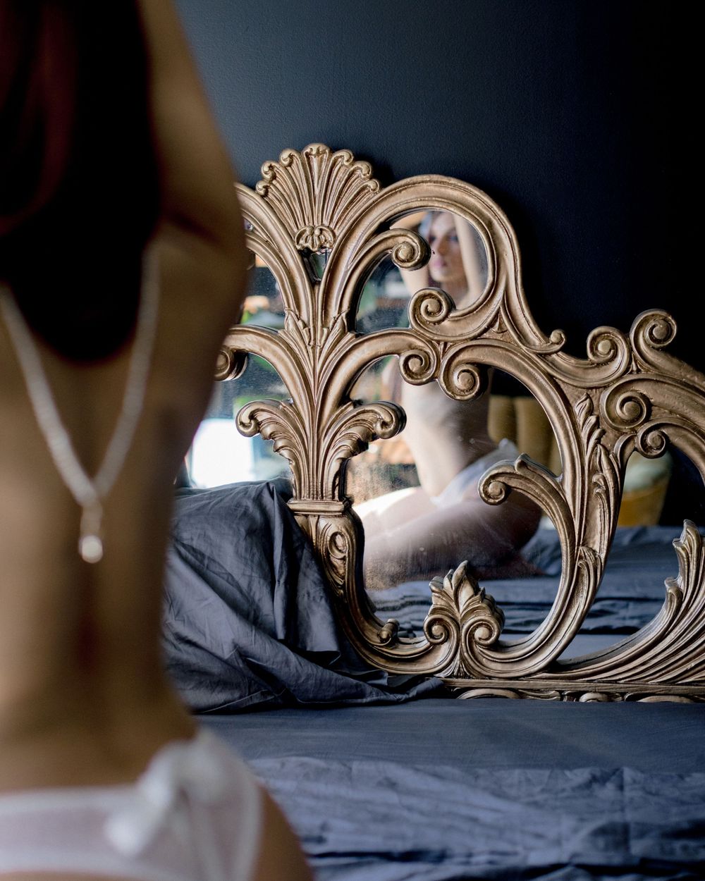 Ornate vintage mirror with decorative scrollwork design reflects a blurred bedroom interior.