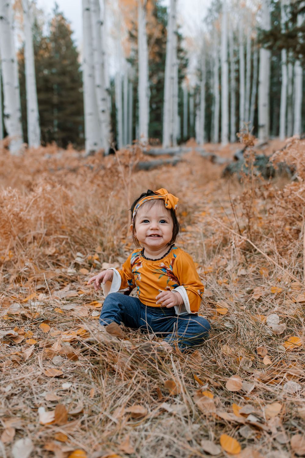Toddler in orange clothing and bow smiles while sitting in fallen autumn leaves.