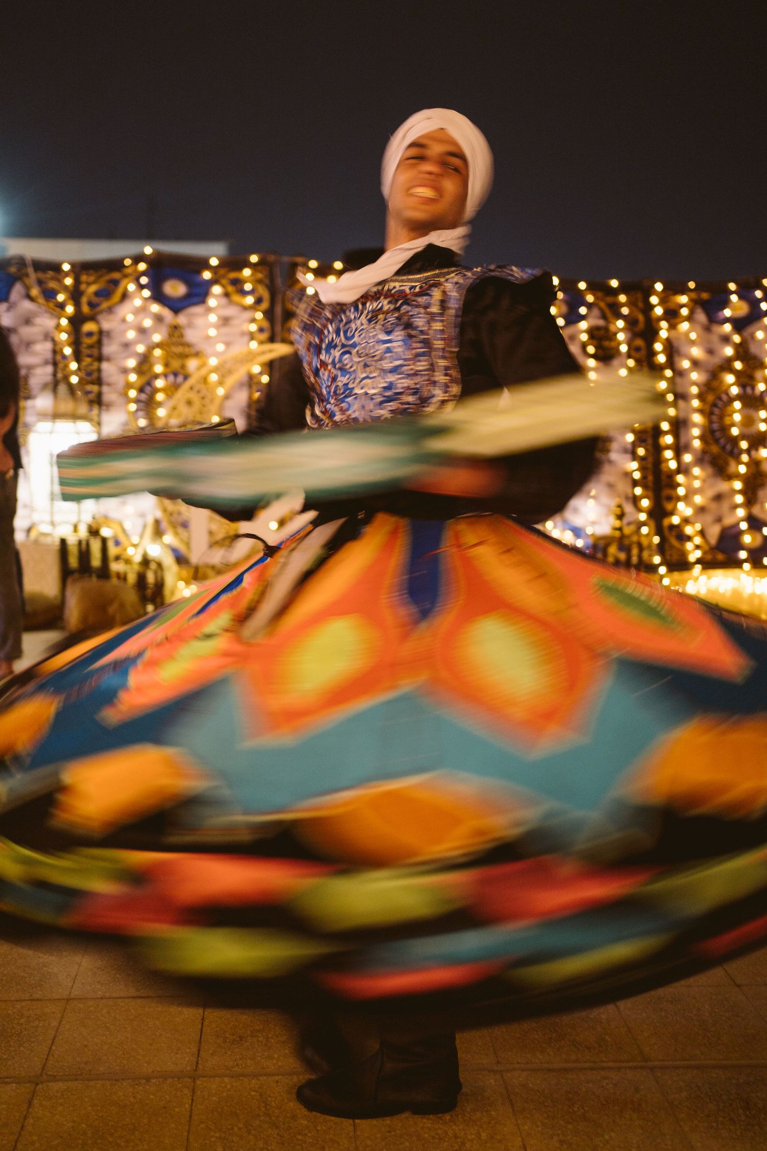 A performer in traditional dress spins in a vibrant swirling costume against a backdrop of glowing holiday lights.
