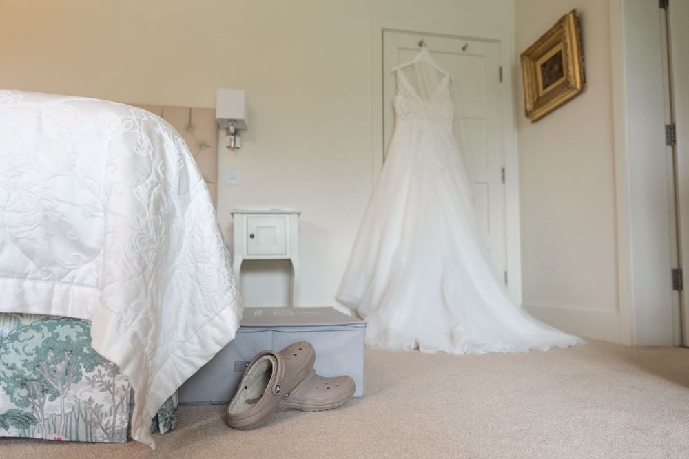 A white wedding dress hangs on display in a bedroom with gray shoes on the floor.