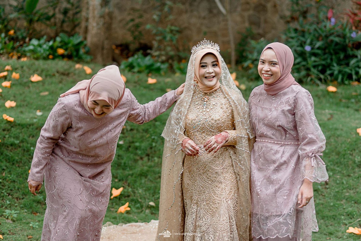 Wedding party members in mauve dresses share laughter with bride in gold dress on garden lawn.