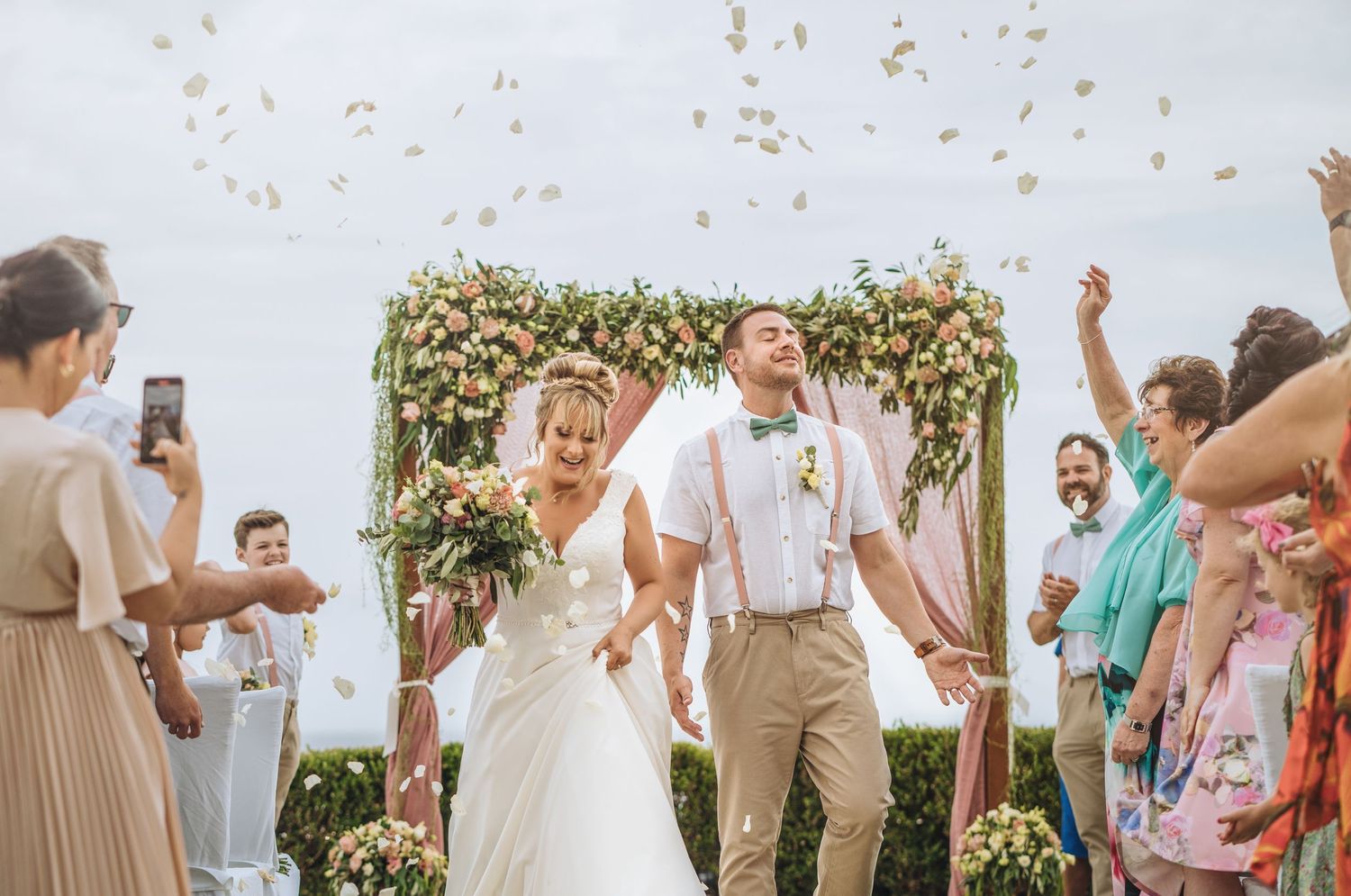 Outdoor wedding celebration with guests throwing confetti against a floral backdrop.