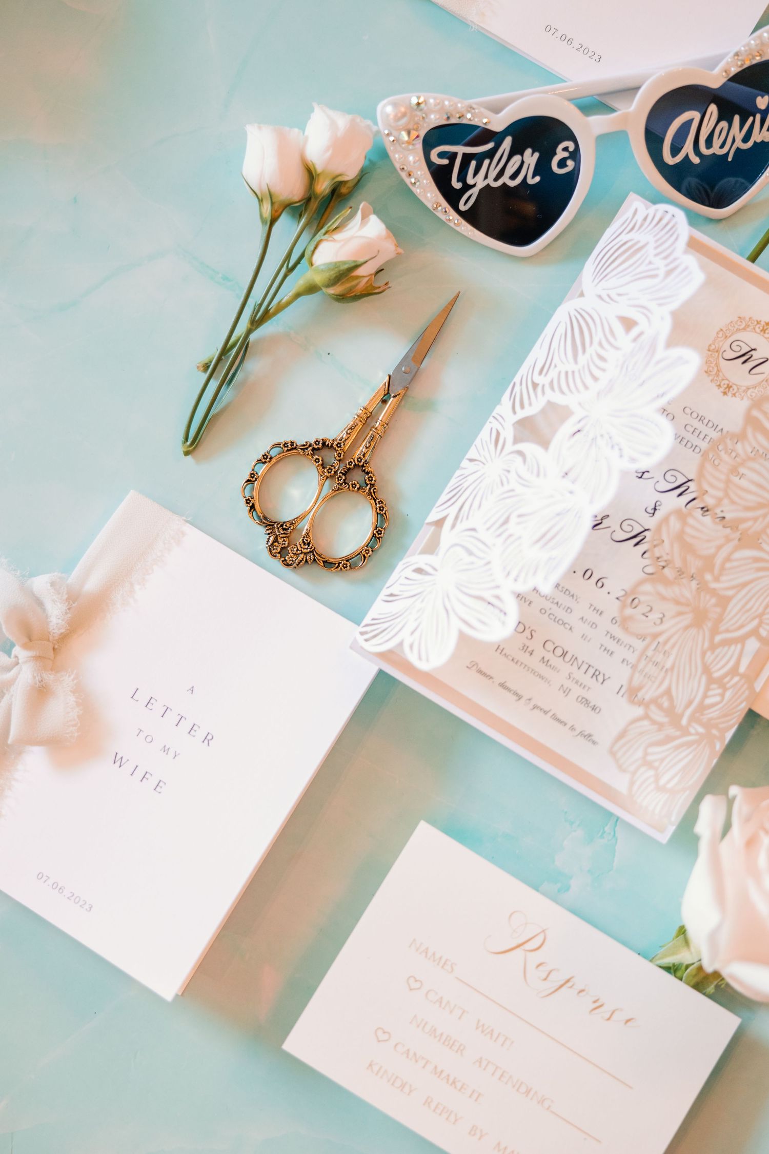 Wedding stationery flatlay with laser-cut invitation, sunglasses, scissors, and roses.