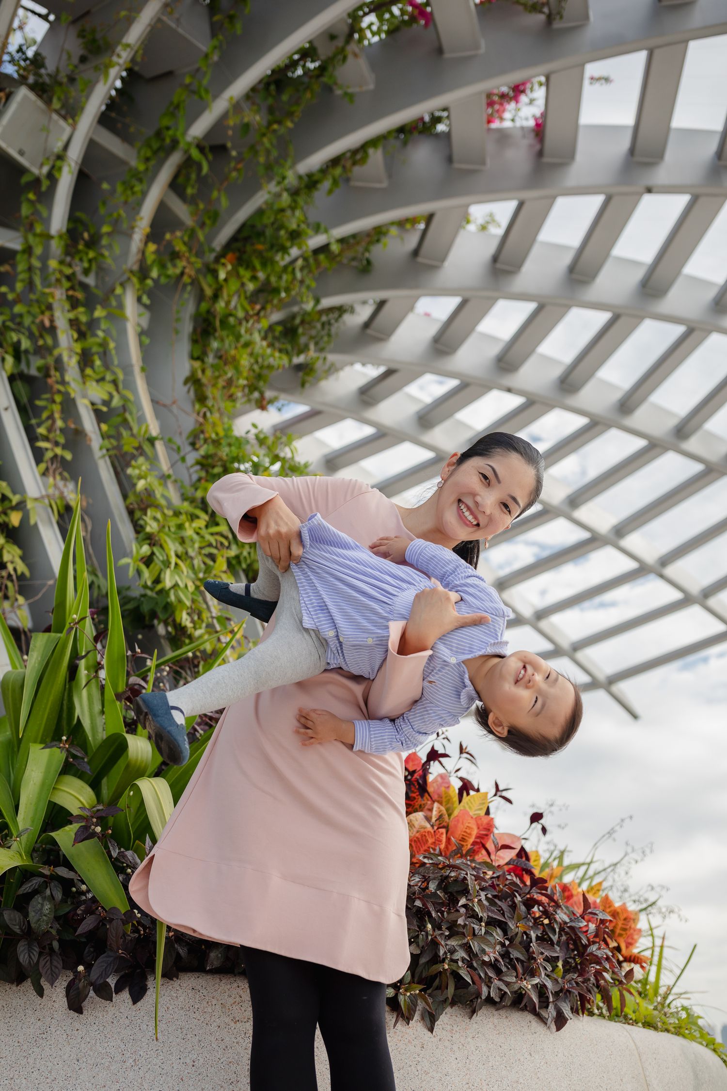 Playful mother and daughter portraits under a white pergola surrounded by tropical plants and flowers.