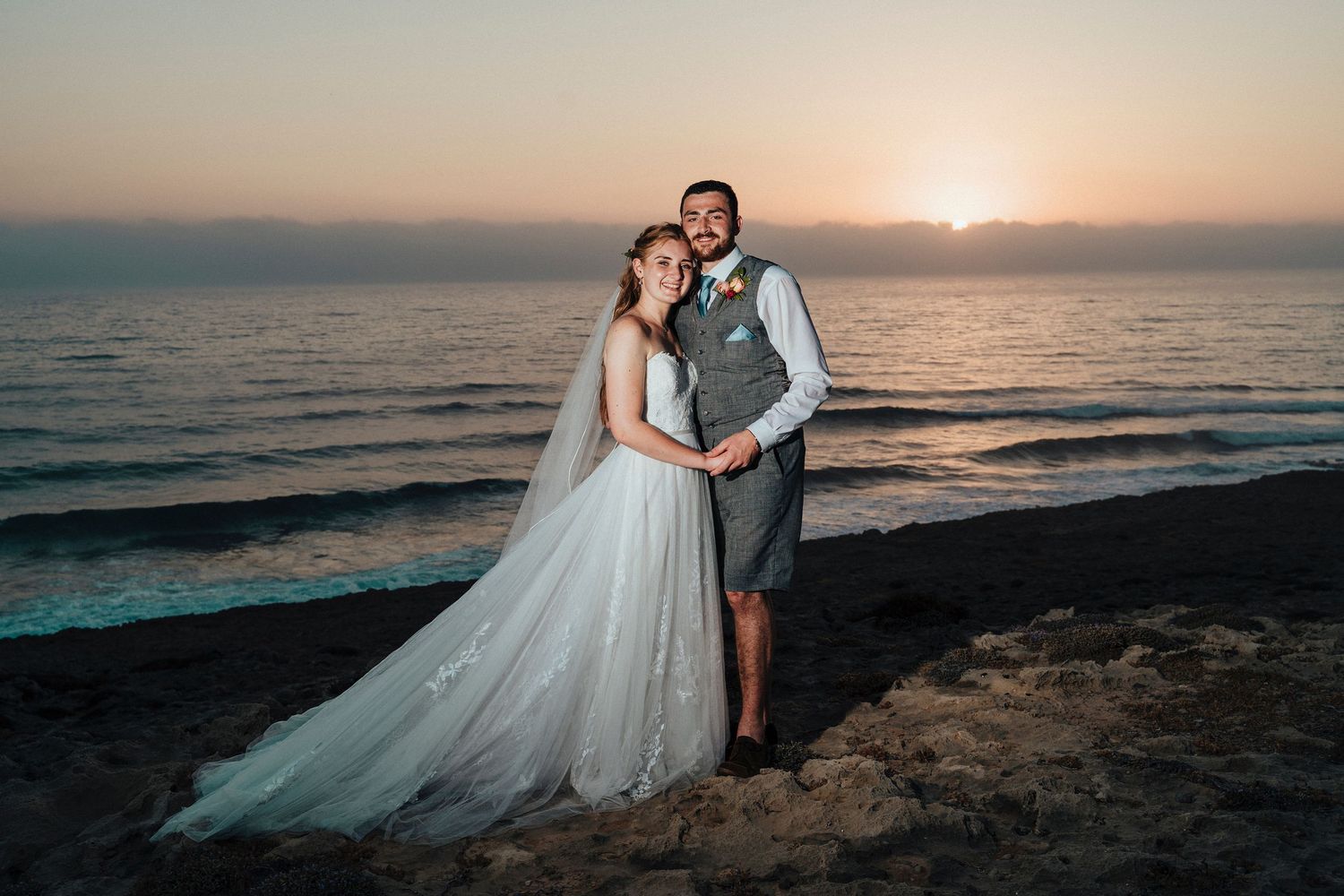 Sunset beach wedding portrait of couple in formal attire against dramatic ocean backdrop.