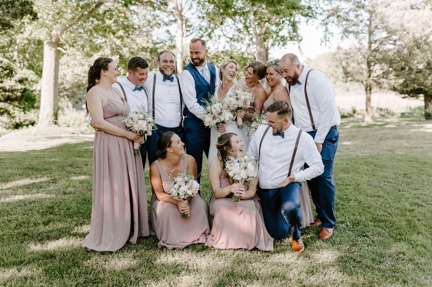 Wedding group poses together on grassy lawn with spring trees in background.